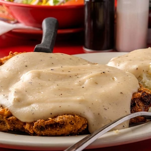 Chicken fried steak with gravy and mashed potatoes on a white plate with a drink and sauce.