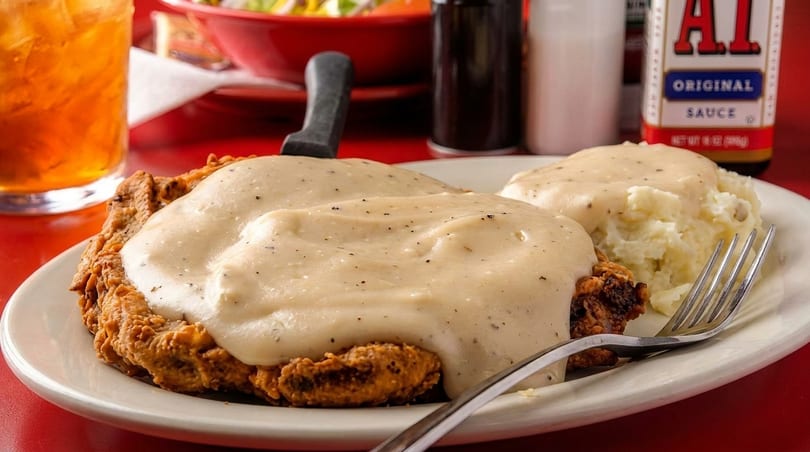 Chicken fried steak with gravy and mashed potatoes on a white plate with a drink and sauce.