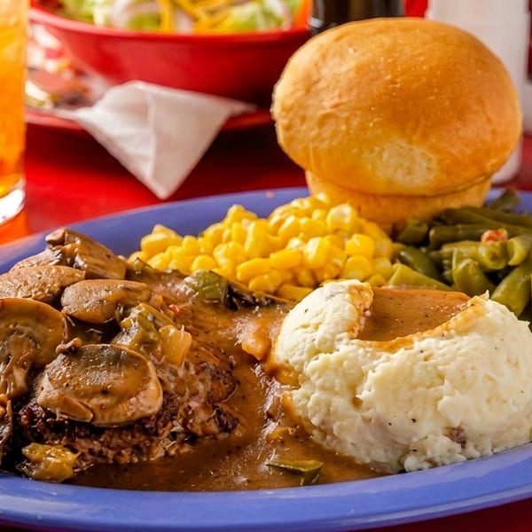 A blue plate with Salisbury steak, mashed potatoes, green beans, corn, and a dinner roll.