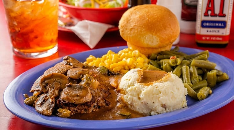 A blue plate with Salisbury steak, mashed potatoes, green beans, corn, and a dinner roll.