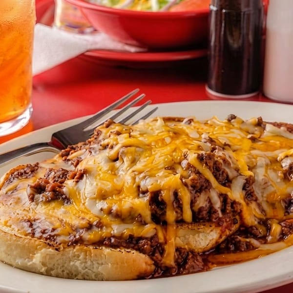 Chili cheese dog on a white plate with a fork, iced tea, and A1 sauce on a red table.