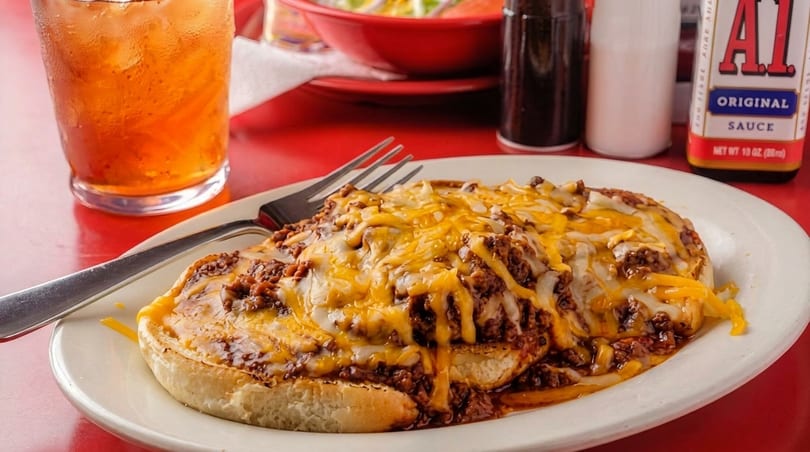 Chili cheese dog on a white plate with a fork, iced tea, and A1 sauce on a red table.