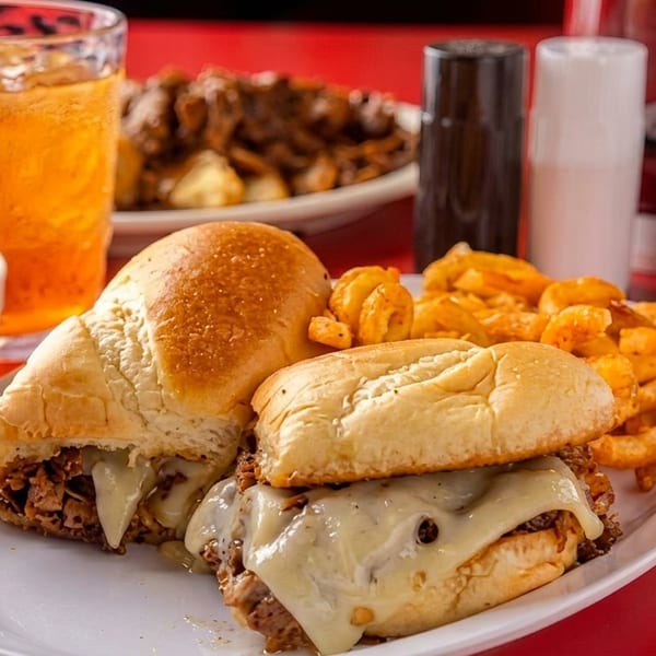 Two cheesesteak sandwiches with curly fries, a drink, and A1 sauce on a red table.